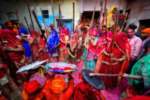 A vibrant scene from the Lathmar Holi festival in Barsana, India, where women dressed in traditional red and blue sarees playfully strike men with sticks as part of the ritual. The men, wearing turbans and holding shields, crouch to protect themselves while the ground is covered in bright pink Holi colors, creating a lively and culturally rich atmosphere.